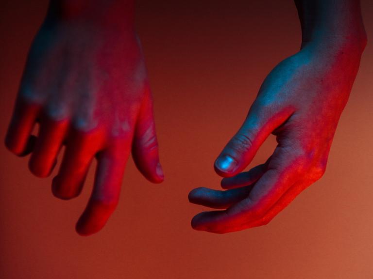 Zwei Hände, die blau, lila und rot beleuchtet sind, hängen von oben herab.