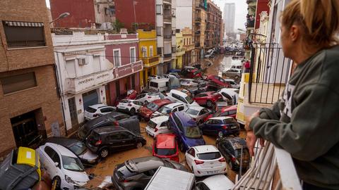 In Valencia blickt eine Frau vom Balkon aus auf eine Straße, in der die inzwischen abgelaufene Überschwemmung Autos wie Spielzeuge zusammen- und teilweis übereinander geschoben hat. 