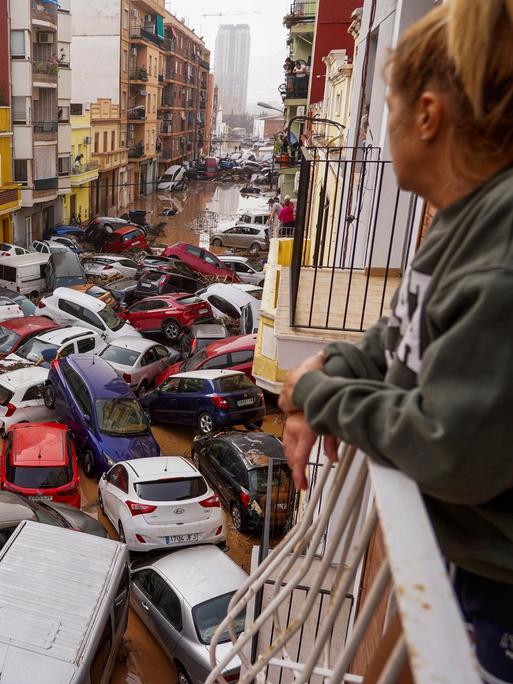 In Valencia blickt eine Frau vom Balkon aus auf eine Straße, in der die inzwischen abgelaufene Überschwemmung Autos wie Spielzeuge zusammen- und teilweis übereinander geschoben hat. 
