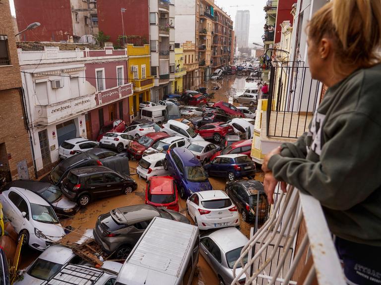 In Valencia blickt eine Frau vom Balkon aus auf eine Straße, in der die inzwischen abgelaufene Überschwemmung Autos wie Spielzeuge zusammen- und teilweis übereinander geschoben hat. 
