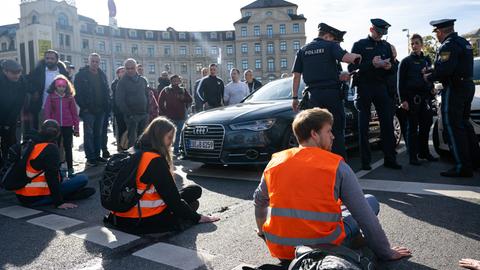 Klimaaktivisten der "Letzten Generation" haben sich am Karlsplatz in der Münchner Innenstadt auf die Fahrbahn geklebt und blockieren die Straße. Sie tragen orangefarbene Signalwesten. In der Näher stehen Polizeibeamte.