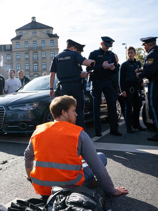 Klimaaktivisten der "Letzten Generation" haben sich am Karlsplatz in der Münchner Innenstadt auf die Fahrbahn geklebt und blockieren die Straße. Sie tragen orangefarbene Signalwesten. In der Näher stehen Polizeibeamte.