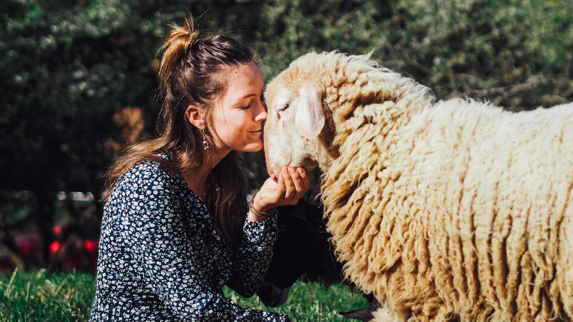 Geraldine Schüle sitzt in einem gepunkteten Kleid auf einer Wiese und küsst ein Schaf.