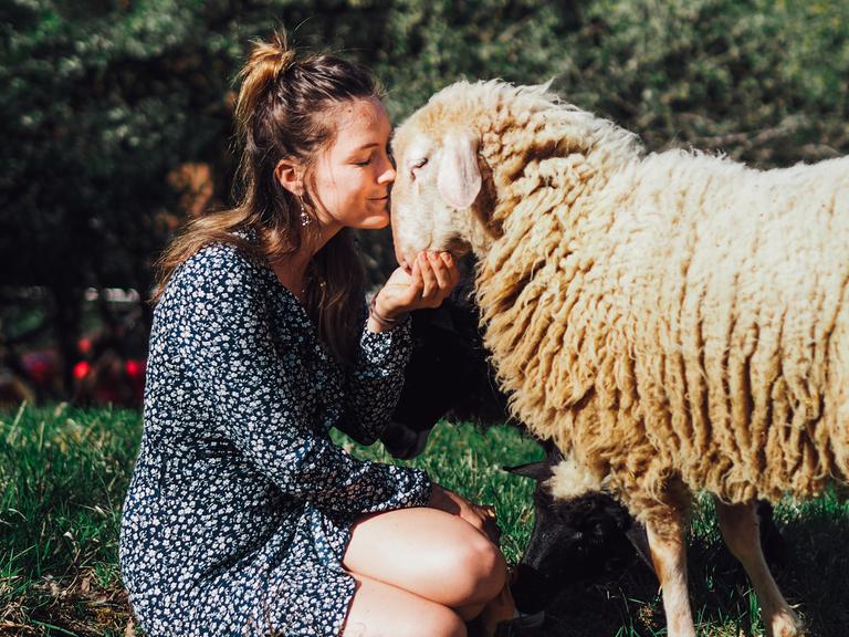 Geraldine Schüle sitzt in einem gepunkteten Kleid auf einer Wiese und küsst ein Schaf.