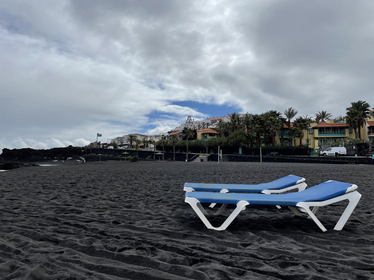 Zwei blaue leere Liegestühle an einem leeren Strand mit schwarzem Sand.