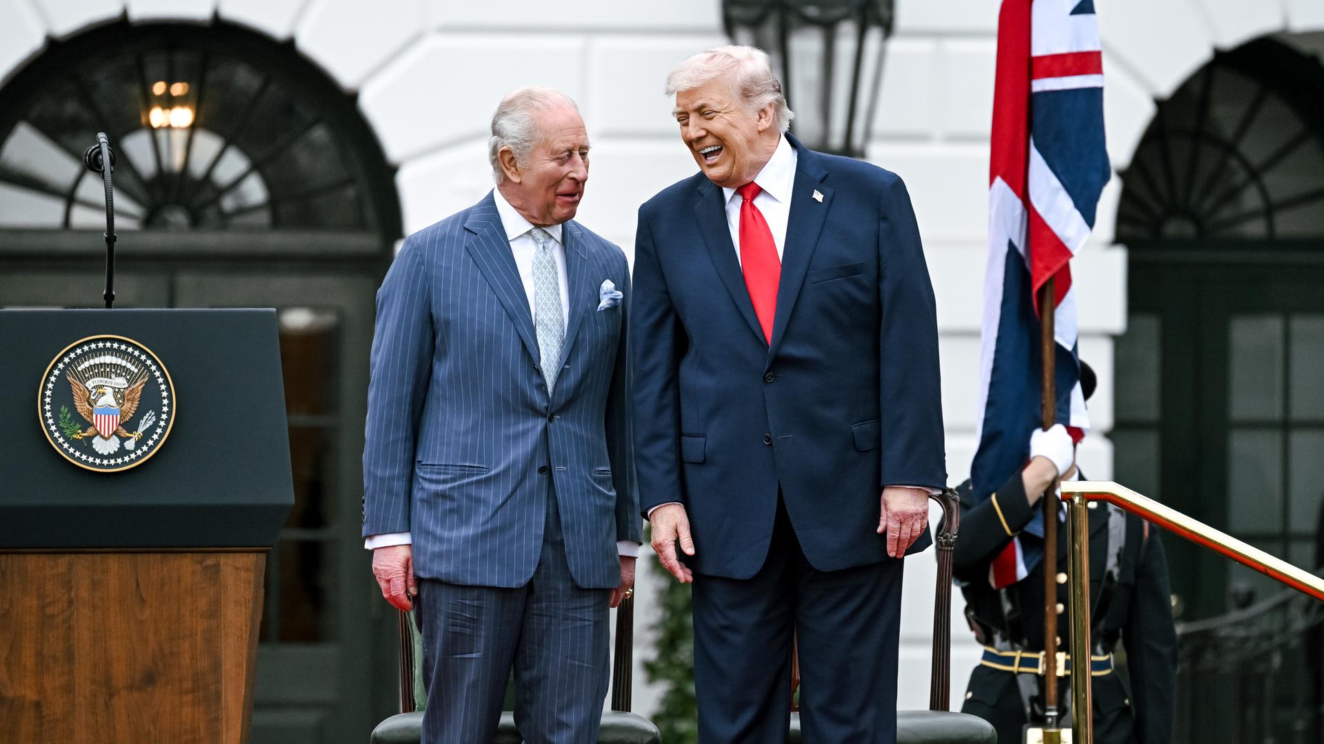 Der britische König Charles III. neben Donald Trump, dem US-Präsidenten. Beide lachen. Sie stehen vor dem Weißen Haus. Beide tragen blaue Anzüge.