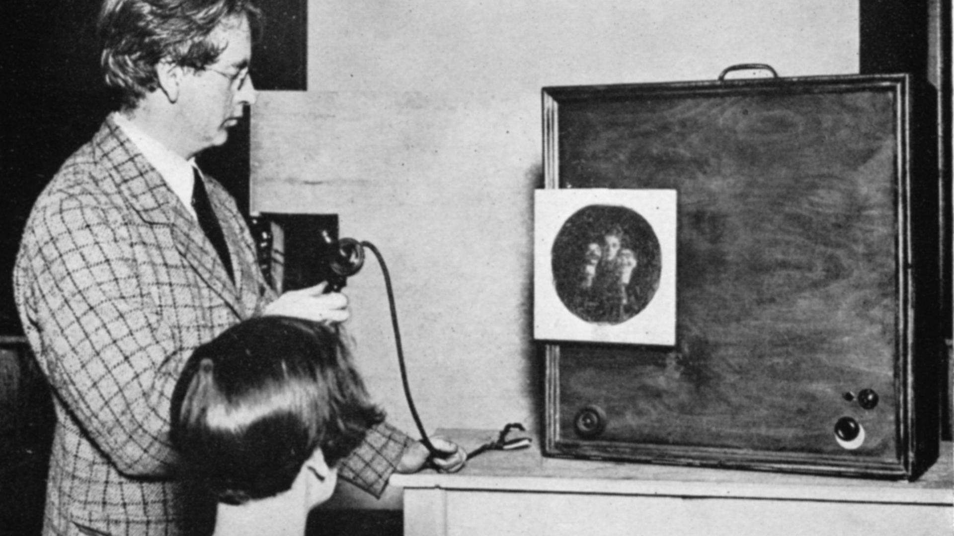 John Baird betrachtet zusammen mit einer Frau, wie ein kreisrundes Bild, das mehrere Gesichter zeigt, auf der Projektionsfläche eines hölzernen Kastens erscheint. John Baird betrachtet zusammen mit einer Frau, wie ein kreisrundes Bild, das mehrere Gesichter zeigt, auf der Projektionsfläche eines hölzernen Kastens erscheint.