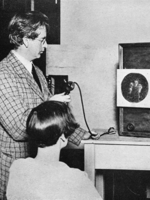 John Baird betrachtet zusammen mit einer Frau, wie ein kreisrundes Bild, das mehrere Gesichter zeigt, auf der Projektionsfläche eines hölzernen Kastens erscheint.