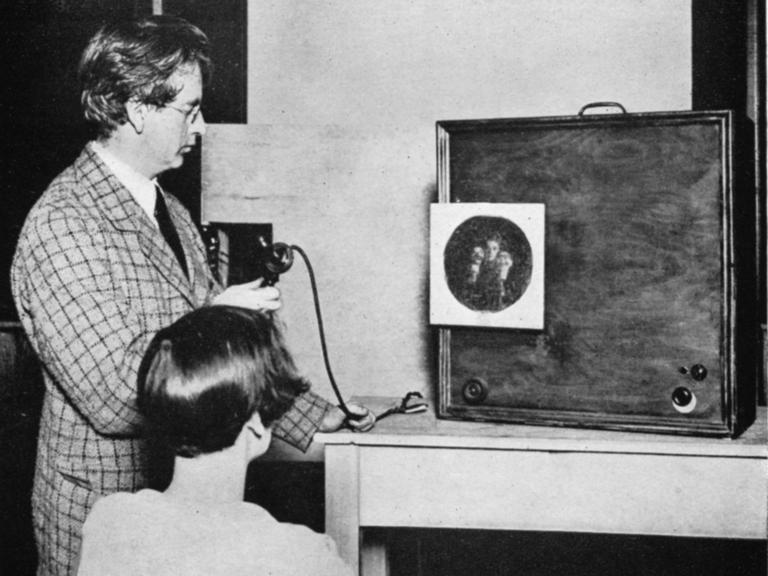 John Baird betrachtet zusammen mit einer Frau, wie ein kreisrundes Bild, das mehrere Gesichter zeigt, auf der Projektionsfläche eines hölzernen Kastens erscheint.