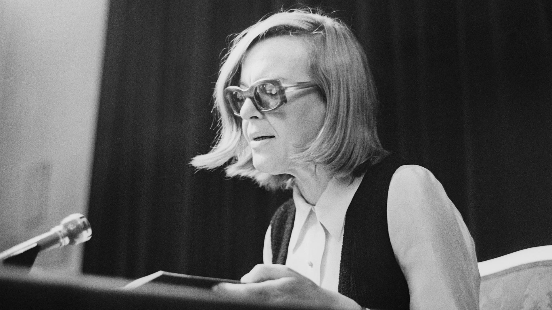 Historisches Schwarzweißfoto von Ingeborg Bachmann bei einer Lesung 1971 in Wien . Sie sitzt sehr aufrecht und trägt eine Brille mit dickem Rahmen.