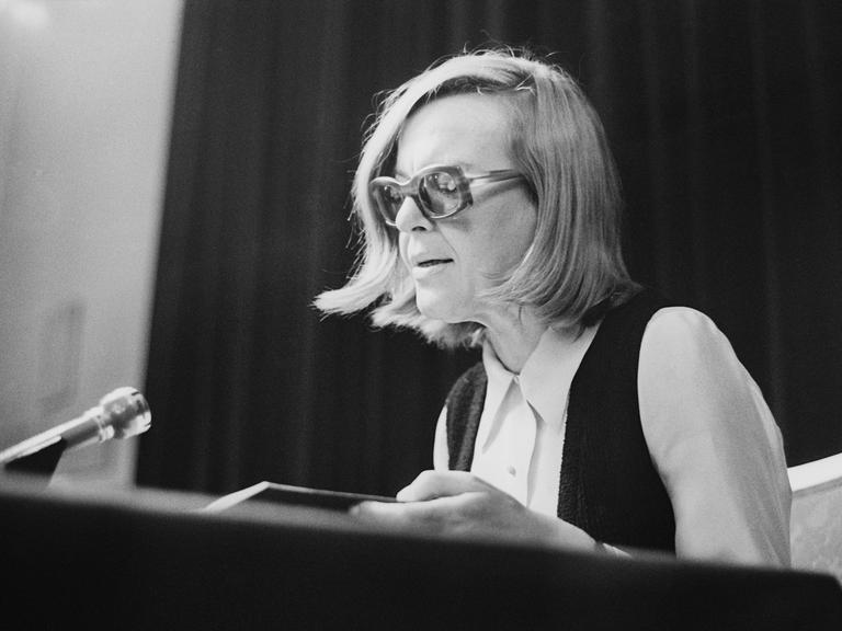 Historisches Schwarzweißfoto von Ingeborg Bachmann bei einer Lesung 1971 in Wien . Sie sitzt sehr aufrecht und trägt eine Brille mit dickem Rahmen.