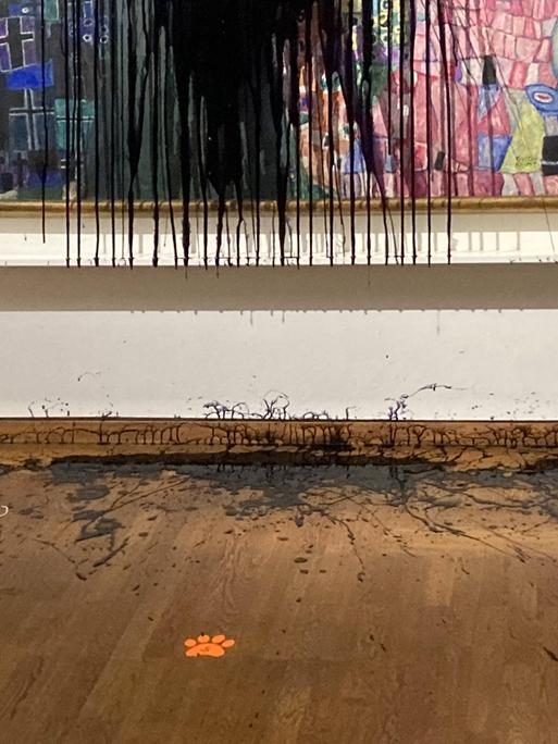 Detail des Gemäldes "Leben und Tod" von Gustav Klimt, was mit schwarzem Öl übergossen ist. Die Wand an der das Gemälde hängt und der Boden des Museums sind ebenfalls mit Öl bedeckt.