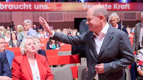 Scholz steht lächelnd vor der ersten Stuhlreihe und winkt jemandem zu. Er ist von der Seite fotografiert.