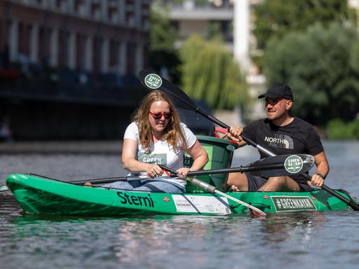Zwei Personen sitzen in einem Kanu auf einem Fluss und holen Müll aus dem Wasser.