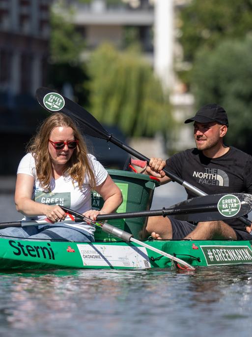 Zwei Personen sitzen in einem Kanu auf einem Fluss und holen Müll aus dem Wasser.