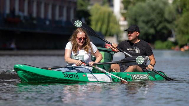 Zwei Personen sitzen in einem Kanu auf einem Fluss und holen Müll aus dem Wasser.