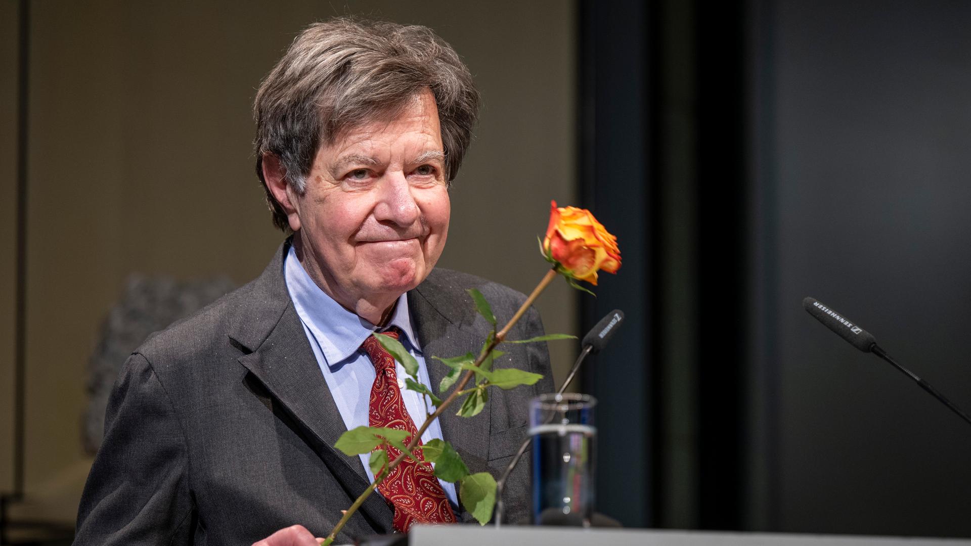 Ein Mann mit Anzug und Krawatte lächelt hinter einem Rednerpult in die Kamera. Er hält eine Rose. Ein Mann mit Anzug und Krawatte lächelt hinter einem Rednerpult in die Kamera. Er hält eine Rose.