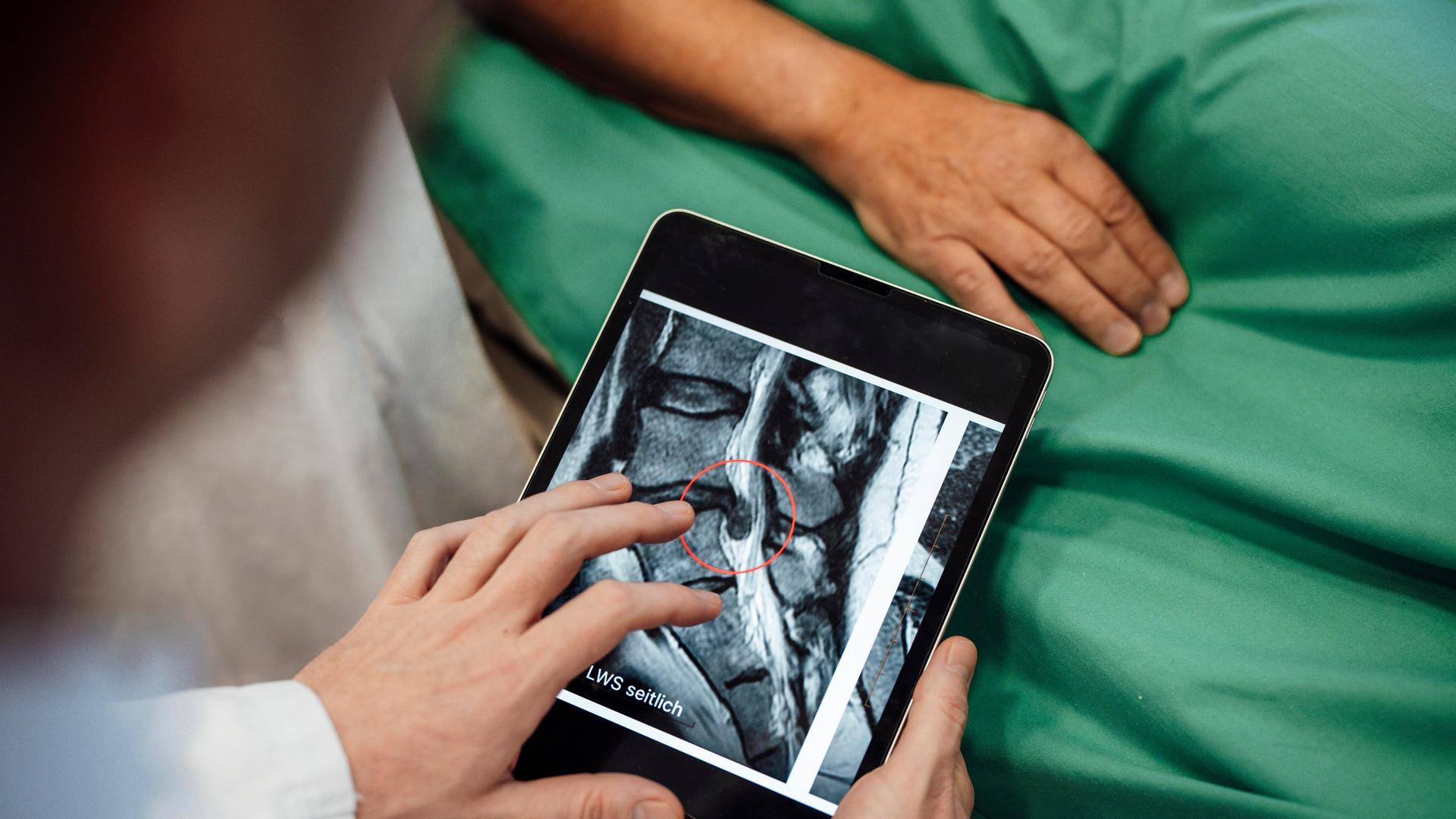 Über die Schulter eines Arzts hinweg ist ein Tablet mit einem Röntgen-Bild zu sehen. Im Hintergrund grüne Krankenhaus-Bettwäsche. Über die Schulter eines Arzts hinweg ist ein Tablet mit einem Röntgen-Bild zu sehen. Im Hintergrund grüne Krankenhaus-Bettwäsche.