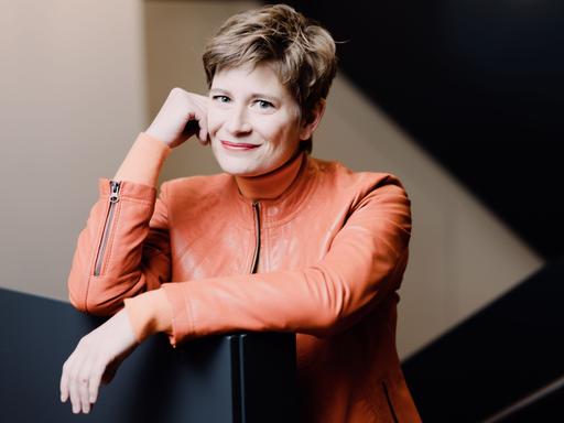 Eine Frau mit kurzem, dunkelblondem Haar steht in einem Treppenhaus, ihren Kopf in die rechte Hand gestützt. Sie trägt eine orangefarbene Lederjacke und lächelt freundlich.