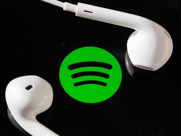 Kopfhörer liegen auf dem Bildschirm eines Smartphones, auf dem das Logo vom Musik-Streaming-Dienst Spotify angezeigt wird.