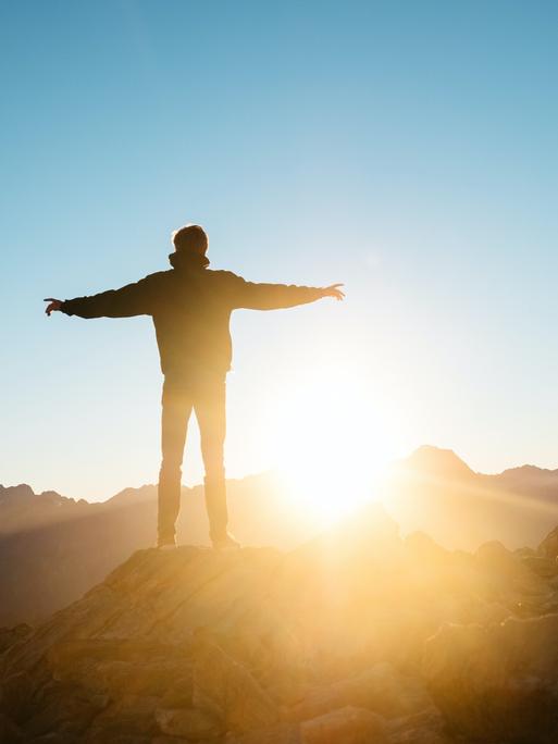 Ein Mann mit ausgebreiteten Armen steht auf einem Berggipfel und schaut der Sonne entgegen.