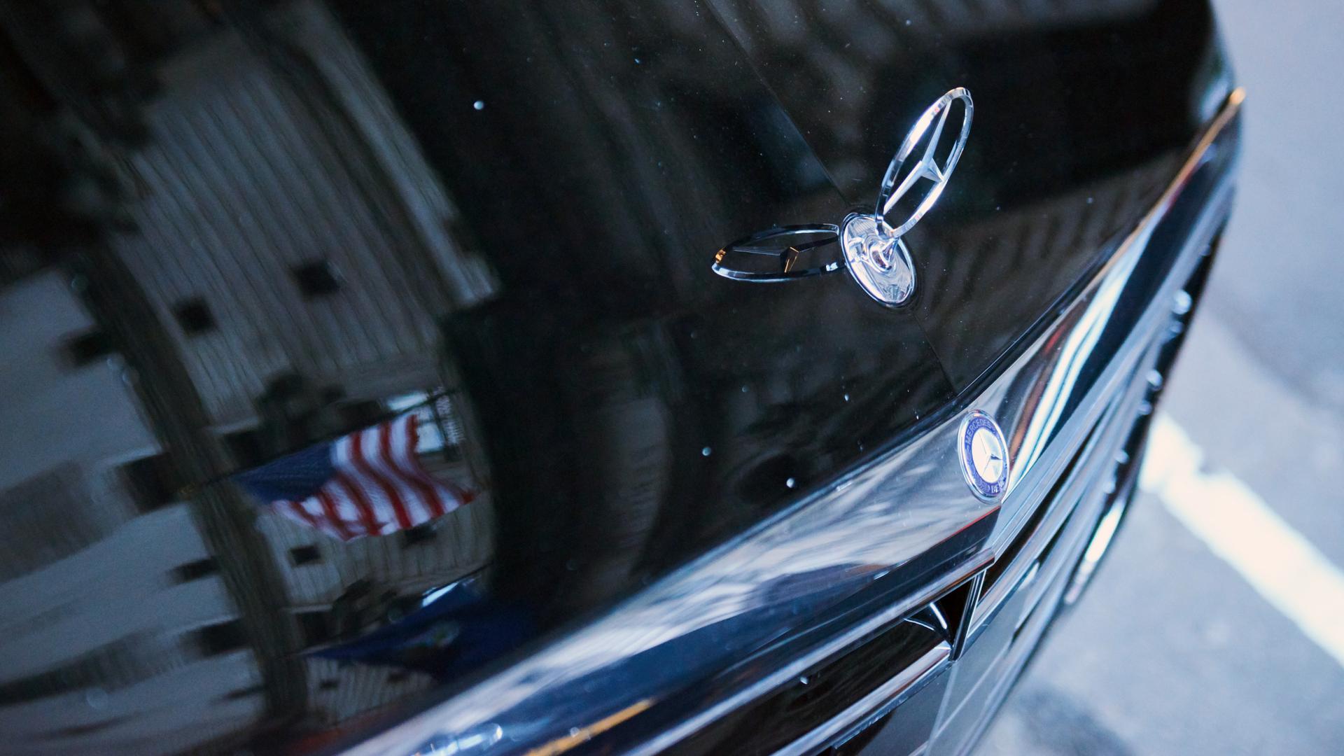 Ein Mercedes-Stern auf einem Schwarzen Mercedes-Auto. Die amerikanische Flagge spiegelt sich auf der Motorhaube. 