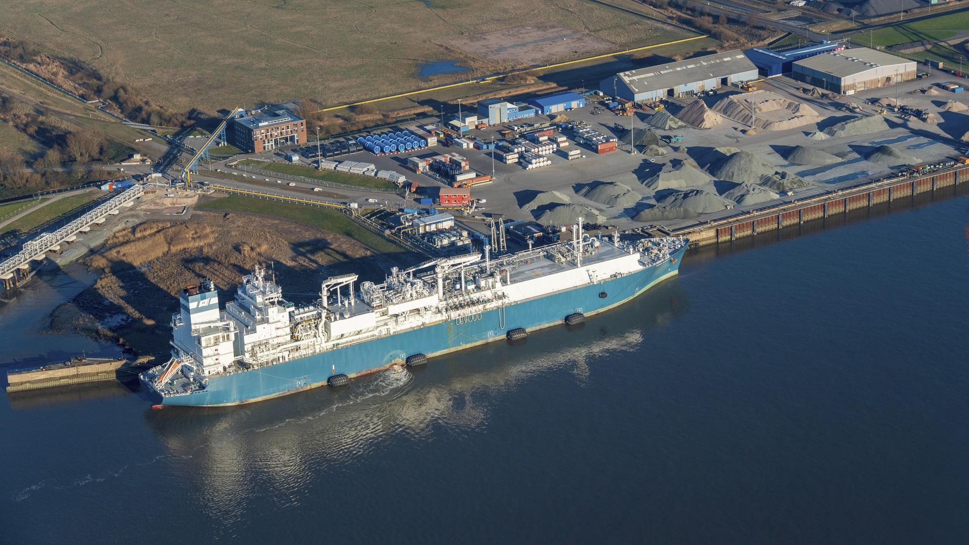 Luftaufnahme vom Elbhafen Brunsbüttel. Links ein blaues Schiff im Wasser, rechts der Hafen mit Containern und Kieshaufen. Luftaufnahme vom Elbhafen Brunsbüttel. Links ein blaues Schiff im Wasser, rechts der Hafen mit Containern und Kieshaufen.