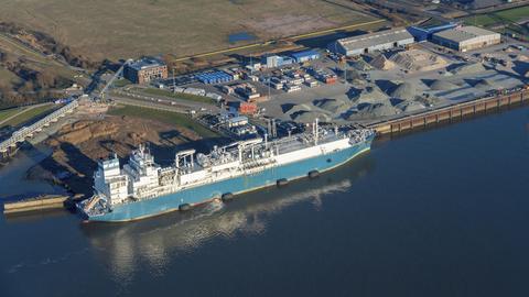 Luftaufnahme vom Elbhafen Brunsbüttel. Links ein blaues Schiff im Wasser, rechts der Hafen mit Containern und Kieshaufen.