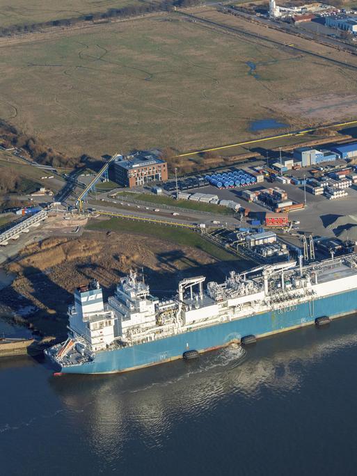 Luftaufnahme vom Elbhafen Brunsbüttel. Links ein blaues Schiff im Wasser, rechts der Hafen mit Containern und Kieshaufen.
