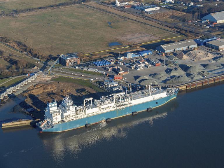 Luftaufnahme vom Elbhafen Brunsbüttel. Links ein blaues Schiff im Wasser, rechts der Hafen mit Containern und Kieshaufen.