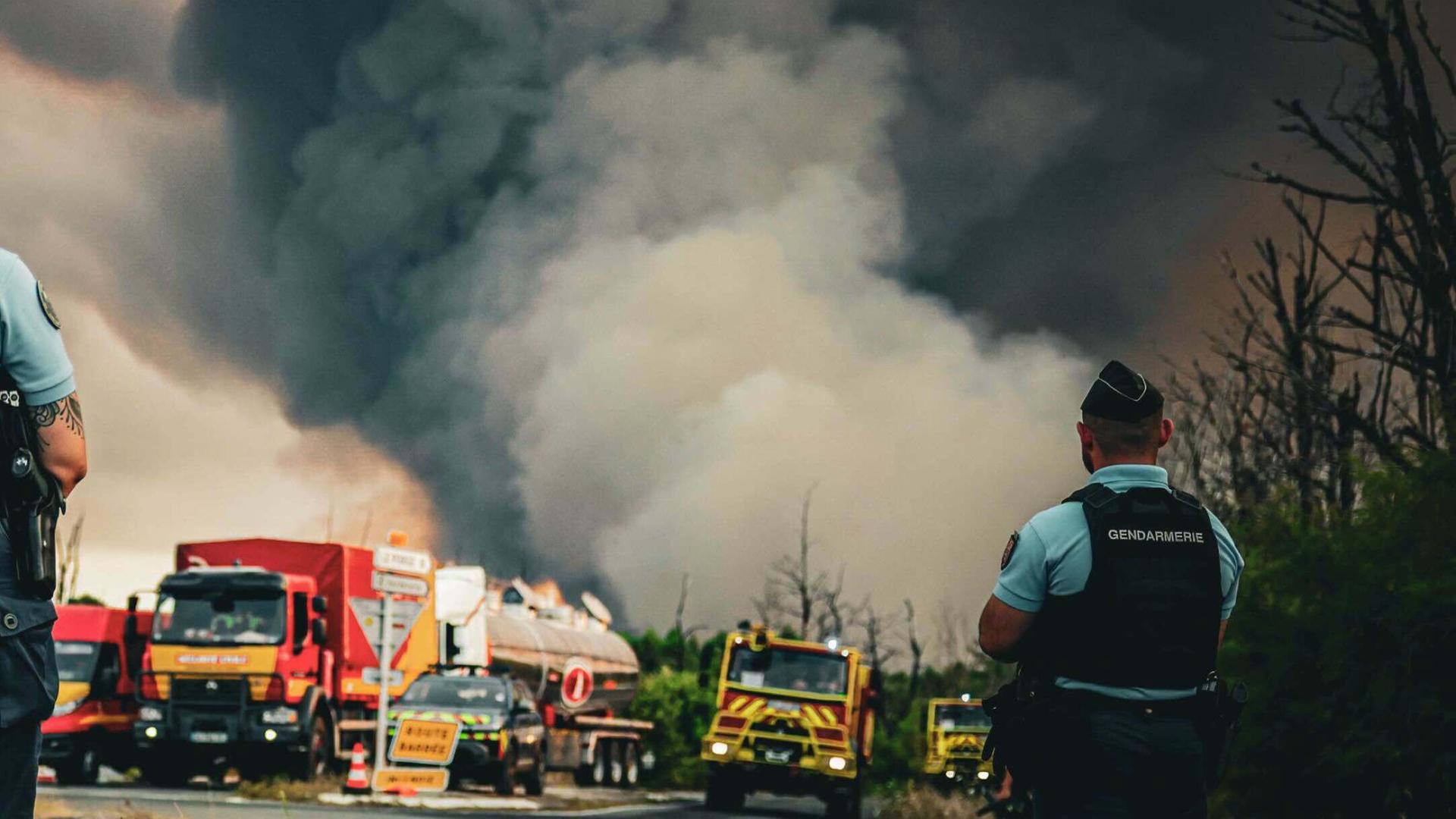 In der Region Gironde wüten Waldbrände. Es steigen dunkle Rauchwolken auf. Davor sind Löschfahrzeuge und ein Polizist auf einer Straße zu sehen. In der Region Gironde wüten Waldbrände. Es steigen dunkle Rauchwolken auf. Davor sind Löschfahrzeuge und ein Polizist auf einer Straße zu sehen.