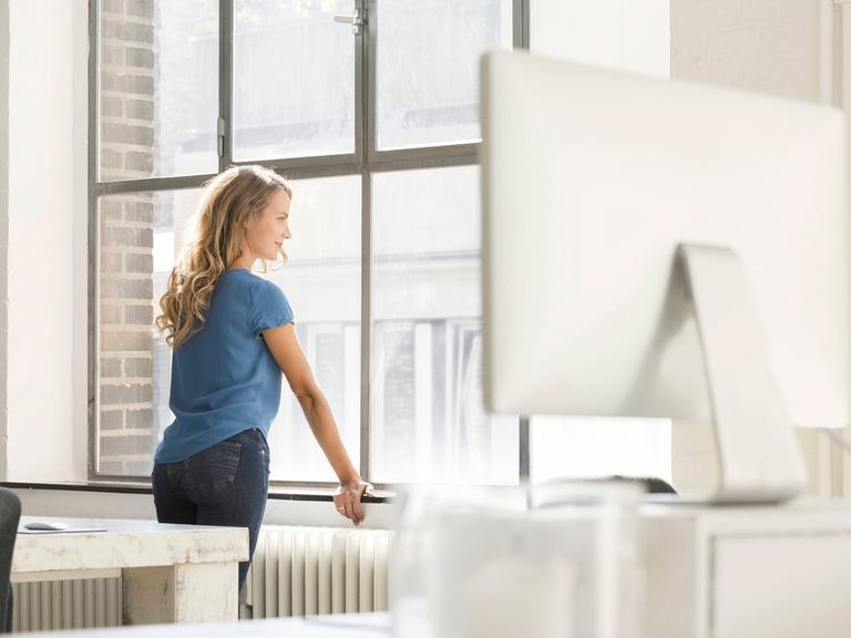 Ein junge blonde Frau mit langen Haaren und hellblauem T-Shirt steht in einem fabriketagen-ähnlichem Büro und schaut aus dem Fenster.