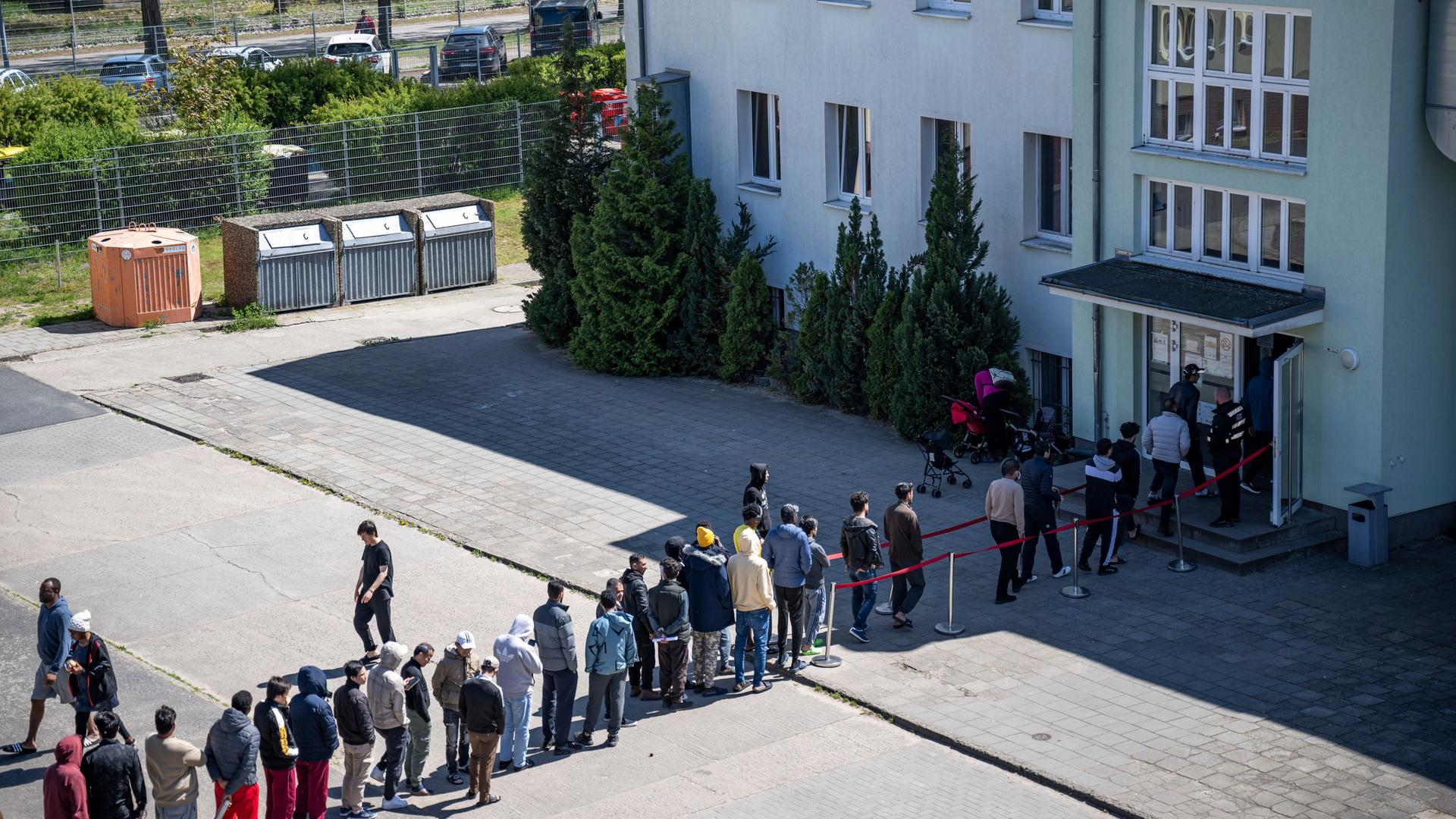 Menschen stehen in einer langen Schlange auf einem Hof vor einem Gebäude und warten auf eine Aufnahme in eine Einrichtung für Geflüchtete.