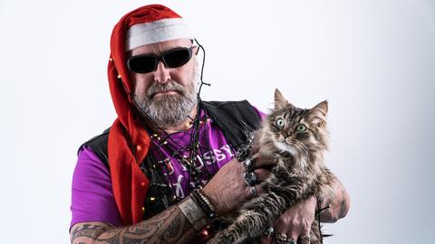 Ein Mann voller Tattoos und Vollbart trägt Weihnachtsmannmütze und Brille, eine Weihnachtsbaumlichterkette um Kopf und Hals gewickelt sowie eine Katze auf dem Arm, die er grault.