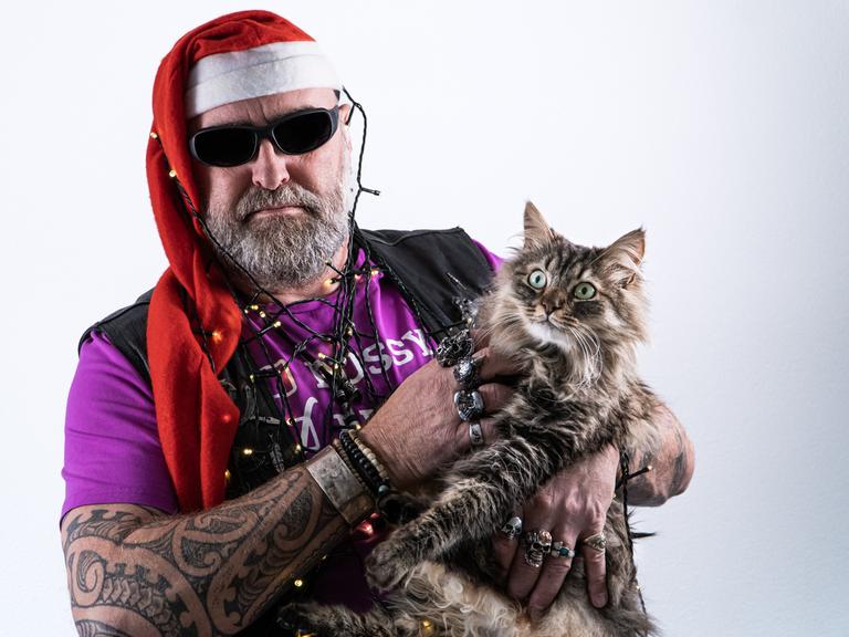 Ein Mann voller Tattoos und Vollbart trägt Weihnachtsmannmütze und Brille, eine Weihnachtsbaumlichterkette um Kopf und Hals gewickelt sowie eine Katze auf dem Arm, die er grault.