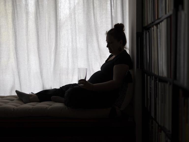 Eine Frau sitzt erschöpft an einem Fenster, in der Hand ein Wasserglas.
