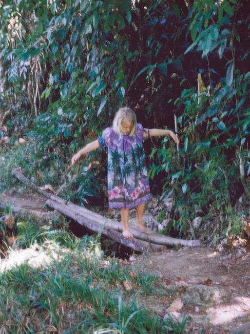 Ein Mädchen namens Elena balanciert im Regenwald auf Baumstämmen über einen kleinen Fluss. Sie trägt ein buntes Kleid und streckt die Arme zur Seite, um das Gleichgewicht zu halten.