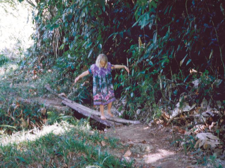 Ein Mädchen namens Elena balanciert im Regenwald auf Baumstämmen über einen kleinen Fluss. Sie trägt ein buntes Kleid und streckt die Arme zur Seite, um das Gleichgewicht zu halten.