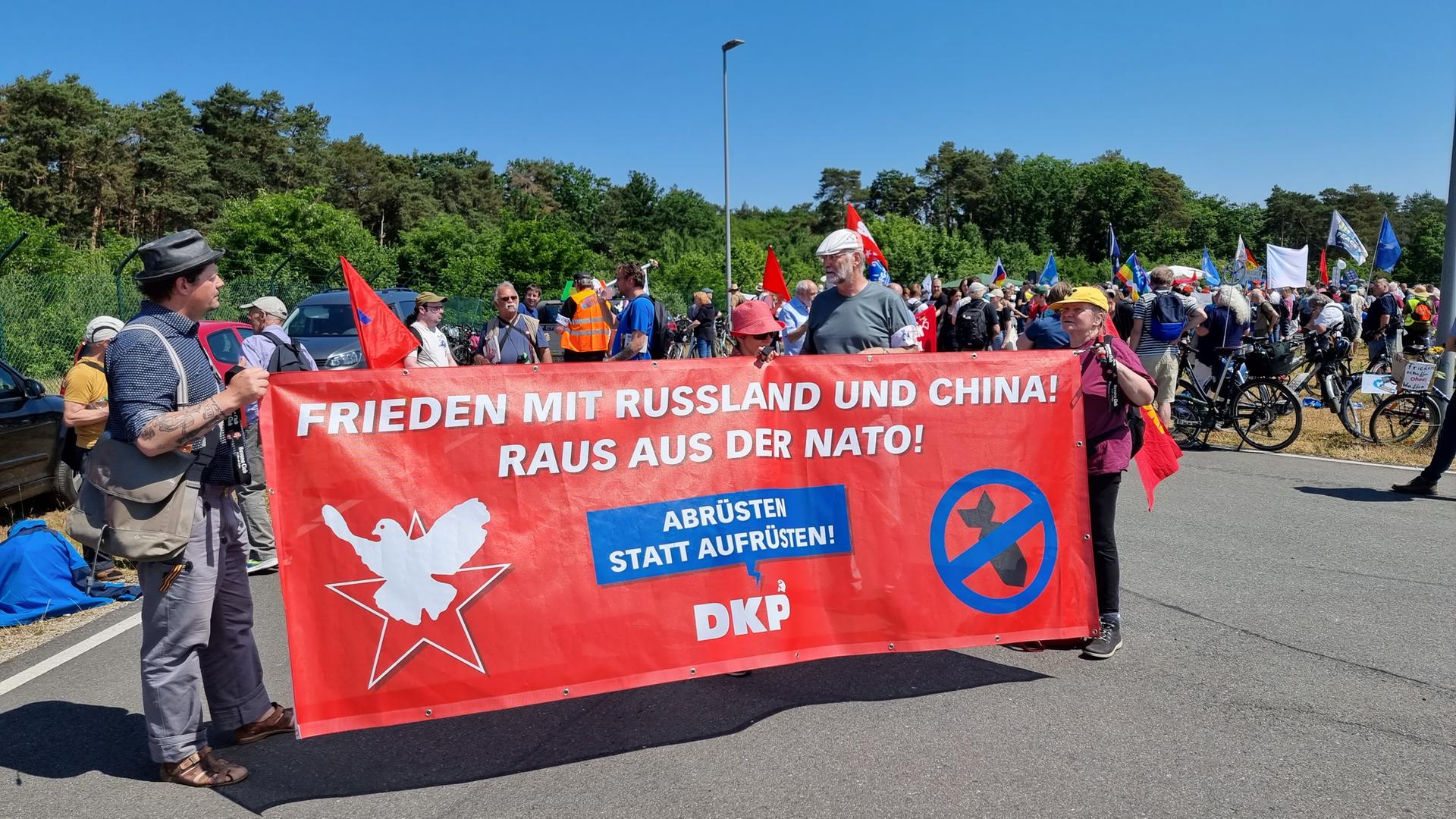 Menschen demonstrieren mit einem Banner mit der Aufschrift "Frieden mit Russland und China! Raus aus der Nato!" vor dem Fliegerhorst in Wunstorf gegen das internationale Luftwaffen-Manöver "Air Defender 2023".