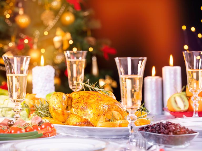 Bild vom reich gedeckten Tisch zu Weihnachten, mit Gänsebraten und Sektgläsern, im Hintergrund verschwommen der Weihnachtsbaum