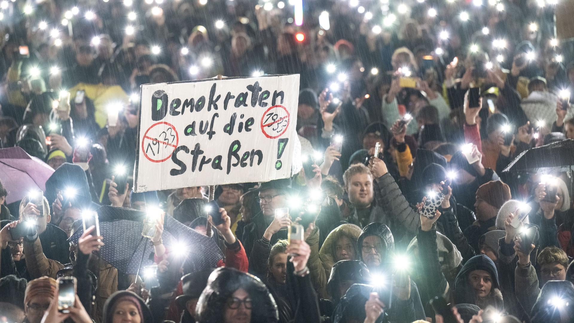 Hessen, Darmstadt: Mehrere Menschen nehmen an einer Demonstration gegen Rechtsextremismus und die AfD teil. Dabei steht auf einem Plakat "Demokraten auf die Straßen". Zum Abschluss der Kundgebung halten die Teilnehmer im strömenden Regen ihre Mobiltelefone in die Höhe.
