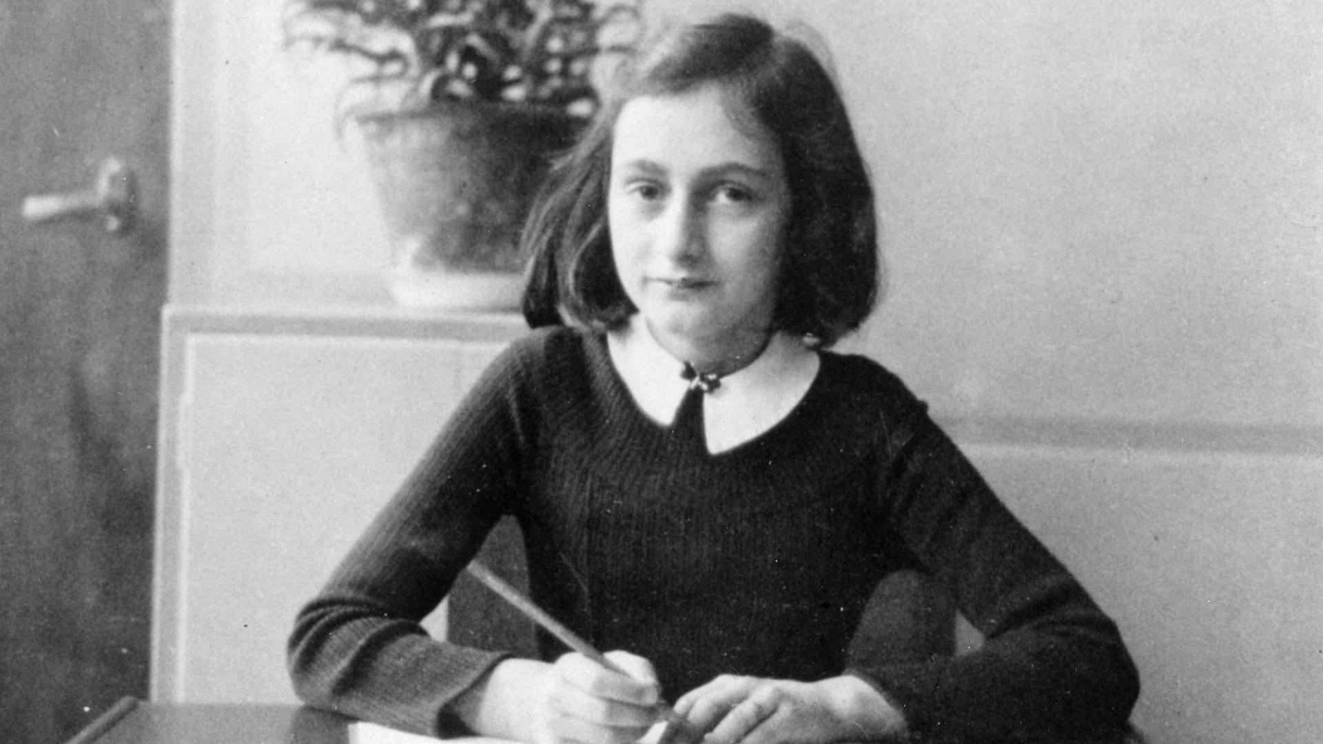 Anne Frank an ihrem Schreibtisch. Sie hat einen Stift in der Hand und ein Heft vor sich und schaut in die Kamera. Anne Frank an ihrem Schreibtisch. Sie hat einen Stift in der Hand und ein Heft vor sich und schaut in die Kamera.