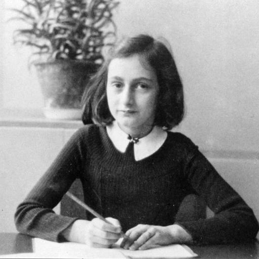 Anne Frank an ihrem Schreibtisch. Sie hat einen Stift in der Hand und ein Heft vor sich und schaut in die Kamera.