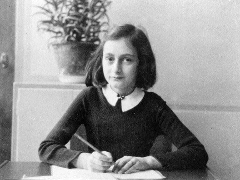 Anne Frank an ihrem Schreibtisch. Sie hat einen Stift in der Hand und ein Heft vor sich und schaut in die Kamera.