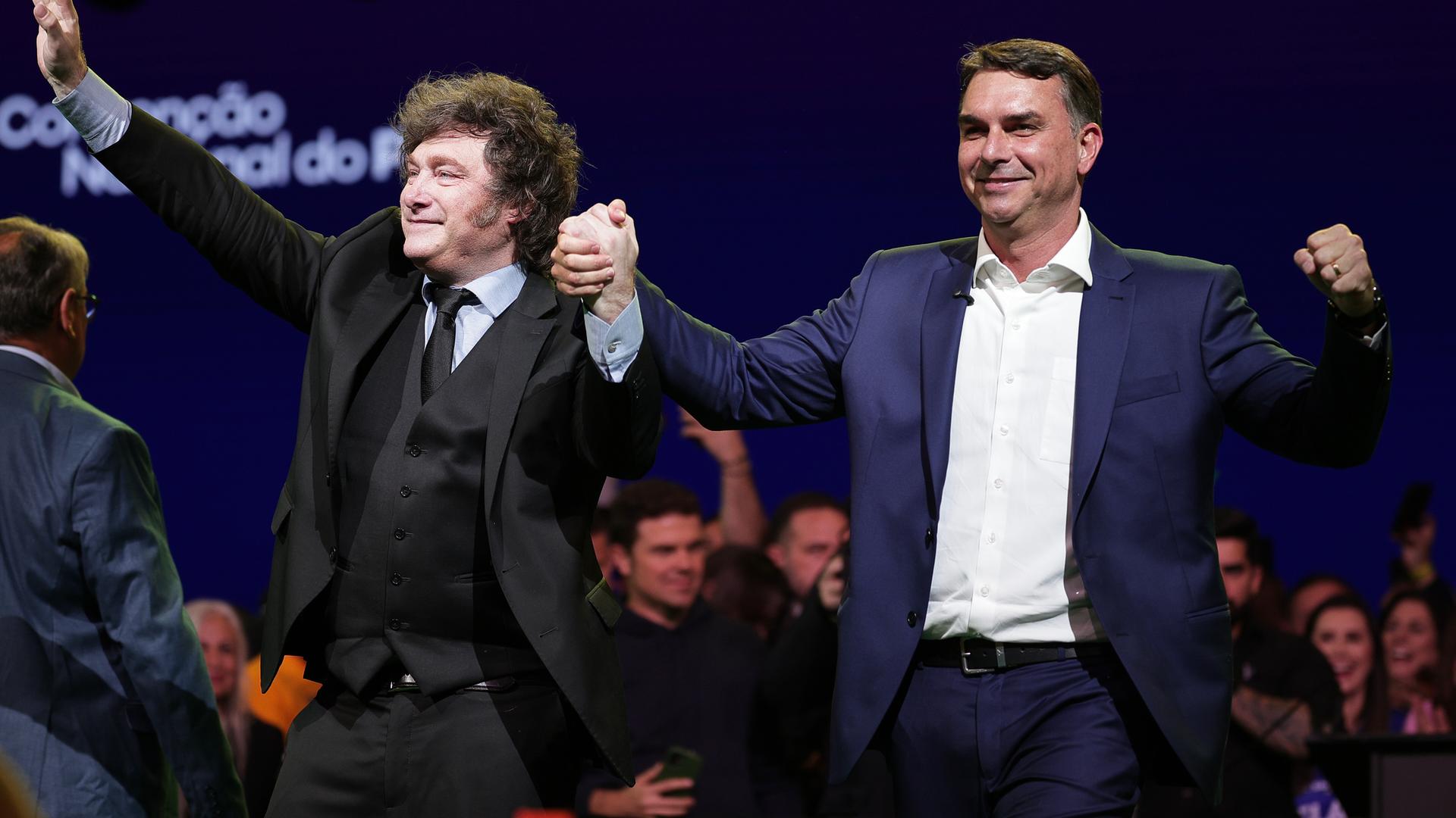 Der brasilianische Senator Flavio Bolsonaro (r.) und Argentiniens Präsident Javier Milei Hand in Hand. Milei winkt mit seiner rechten Hand, Bolsonaro hat seine linke Hand zur Jubelfaust geballt. 