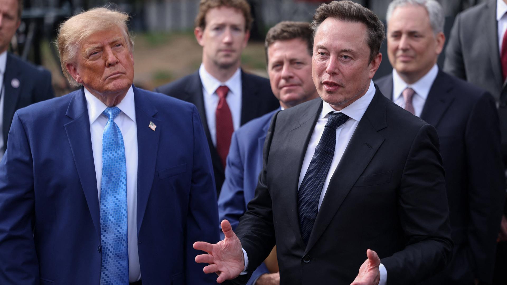 US-Präsident Donald Trump steht links neben Elon Musk, im Hintergrund sind weitere CEOs zu sehen, wie Meta-Chef Mark Zuckerberg