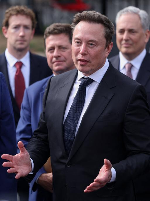 US-Präsident Donald Trump steht links neben Elon Musk, im Hintergrund sind weitere CEOs zu sehen, wie Meta-Chef Mark Zuckerberg