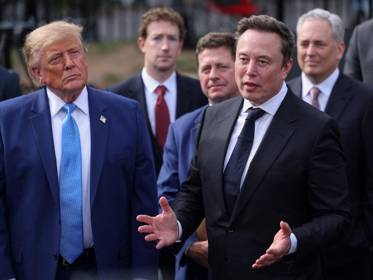 US-Präsident Donald Trump steht links neben Elon Musk, im Hintergrund sind weitere CEOs zu sehen, wie Meta-Chef Mark Zuckerberg