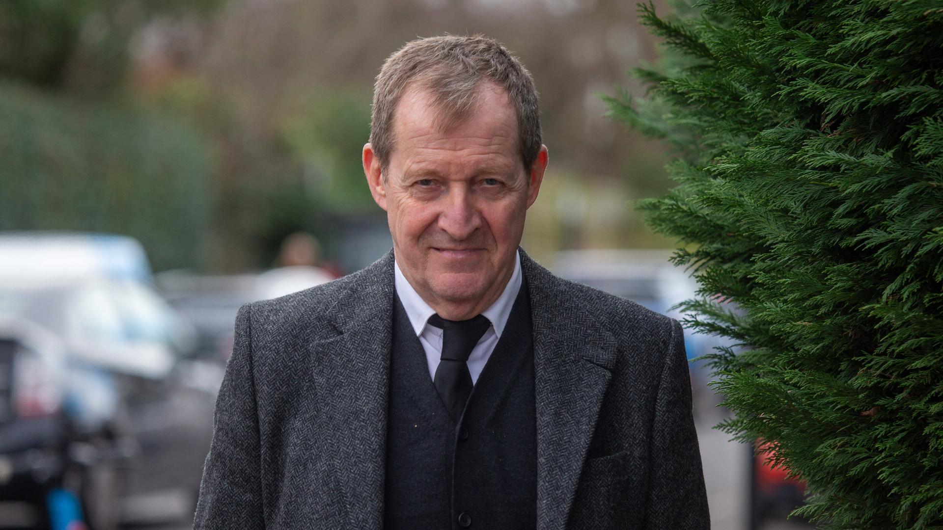 Alastair Campbell vor der St. Mary The Virgin Church in London. Er trägt einen grauen Anzug, einen dunklen Cardigan, ein weißes Hemd und eine schwarze Krawatte. Im Hintergrund sind Büsche und Autos zu erkennen.