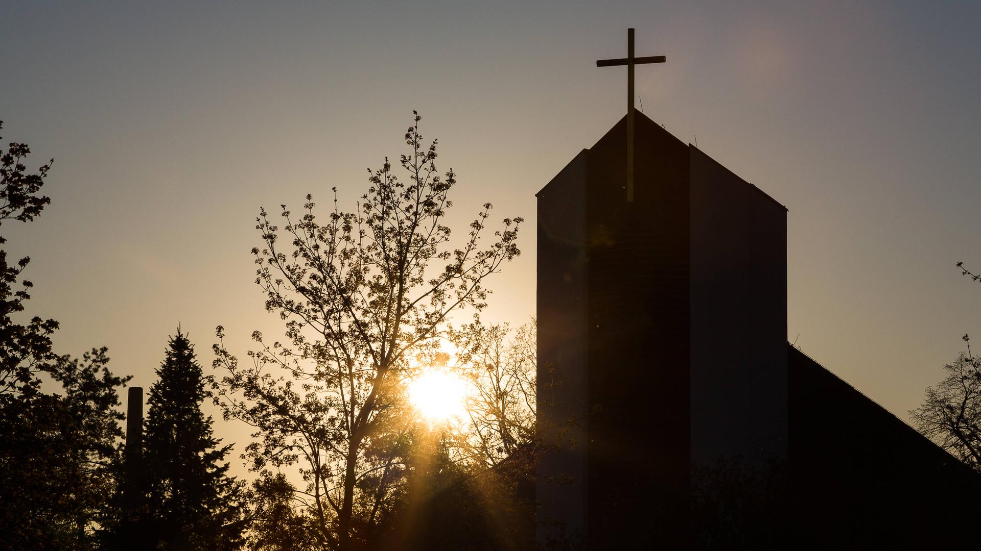 Ein Kirchturm mit Kreuz steht im Gegenlicht der Sonne.